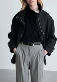 Veste en laine noire mélangée avec un col zippé, associée à un pull noir et un pantalon plissé gris, avec une ceinture noire.