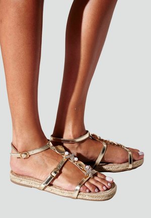 Pieds portant des sandales métalliques dorées avec des décorations de coquillages, d'étoiles de mer et de perles, et des brides à boucle à la cheville sur des semelles en corde beige.