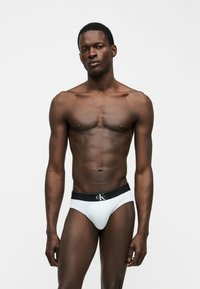 Μυώδης άνδρας στέκεται σε λευκό φόντο φορώντας λευκά εσώρουχα Calvin Klein με μαύρη μέση και λογότυπο στο κέντρο μπροστά.