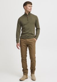 Pull léger tricoté olive à demi-zip ; chinos beiges, coupe ajustée ; bottes chelsea beiges ; texture lisse ; accessoires minimaux ; pose debout décontractée.