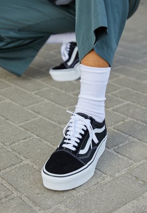 Osoba nosząca czarno-białe sneakersy na platformie, białe skarpetki i zielone, skrócone spodnie, stojąca na szarej ceglanej nawierzchni.