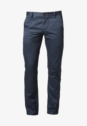 Homme portant un pull à motifs noir, blanc et rouge avec un pantalon bleu, des chaussures beiges et portant un sac noir avec des poignées marron.