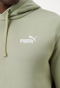 Mand iført en lys grøn hættetrøje med hvidt Puma-logo på brystet, nærbillede af overkroppen og snore.