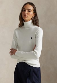 Mujer con pelo largo y castaño que lleva un suéter blanco de cuello alto con un logo azul marino y pantalones oscuros plisados, con los brazos cruzados sobre un fondo neutro.
