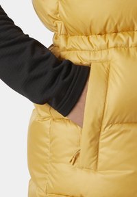 Žlutá nafukovací vesta s hladkým povrchem, se zipovou kapsou a elastickými manžetami, kombinovaná s černým tričkem s dlouhým rukávem pod ní.