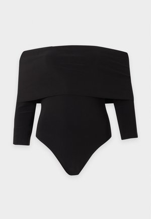 Femme aux longs cheveux foncés portant une mini-robe noire moulante épaules dénudées et des sandales noires à talons hauts, debout avec une main sur la hanche.