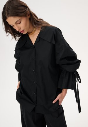 Kobieta o długich brązowych włosach, ubrana w oversize'ową czarną koszulę na guziki z szerokimi mankietami zawiązanymi na nadgarstkach oraz złoty łańcuszek, z rękami w kieszeniach.