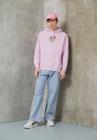 Rozā kapuce ar grafisku dizainu, gaiši zili plata kāju džinsi un balti sporta apavi. Fons ir gluda pelēka siena.