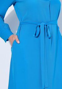 Sininen vyöllinen mekko pitkillä hihoilla, sileällä pinnalla ja sivutaskulla. Vyö on sidottu vyötärölle ja siinä on yksinkertainen muotoilu.