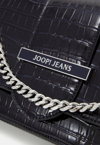 Sac à main noir brillant en simili crocodile avec rabat, chaîne en métal argenté et plaque métallique portant l'inscription "JOOP! JEANS" en lettres blanches.