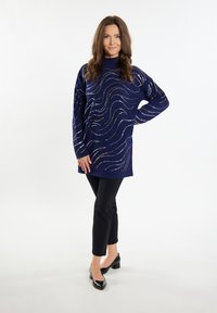 Marineblauer Pullover mit wellenförmigen metallischen Akzenten, langen Ärmeln und hohem Kragen, kombiniert mit schmalen schwarzen Hosen und schwarzen High Heels.