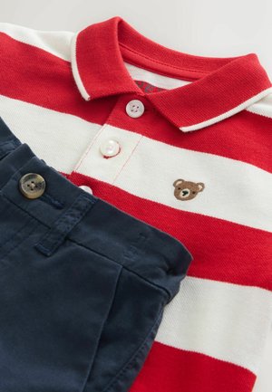 Červeno-bílé pruhované polo tričko s výšivkou medvídka, doplněné tmavě modrými kalhotami s knoflíkovým zapínáním.