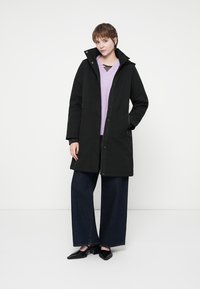 Sort vandafvisende frakke med hætte, knaplukning og sidelommer. Bæres over en lys lilla sweater og brede jeans. Flade sko.