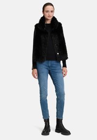 Schwarze Fellweste mit hohem Kragen und Knopfverschluss, getragen über einem schwarzen Oberteil, kombiniert mit hellblauen Jeans und schwarzen Ankle-Boots.