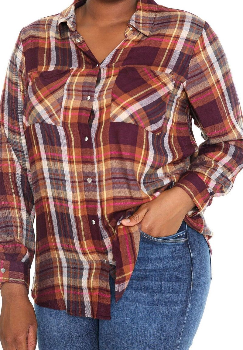 Femme portant une chemise à carreaux boutonnée à manches longues dans des tons bordeaux, orange et jaune, une main dans la poche d'un jean bleu.