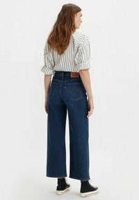 Vaqueros de pierna ancha de color azul oscuro con cintura alta, combinados con una camisa a rayas blanca y negra. Los vaqueros tienen un parche de cuero con el logotipo en la parte posterior.