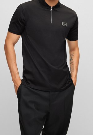 Man draagt een zwart poloshirt met korte mouwen, voorrits en logo patch, zwarte broek, handen in de zakken en tatoeages op zijn rechteronderarm.