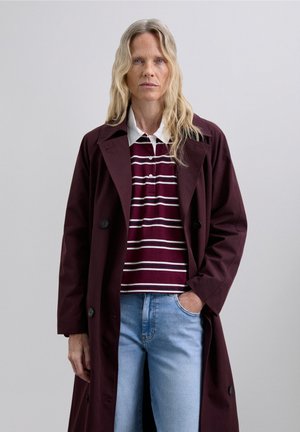Mujer con cabello largo rubio, vistiendo un abrigo burdeos, una camiseta de rayas y jeans azul claro, de pie con una mano en el bolsillo.