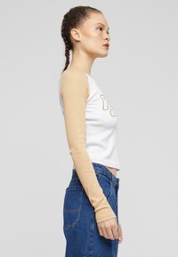 Hvit crop-top med beige lange ermer og trykkede bokstaver. Bært med høy midje blå jeans, som viser en tettsittende design.