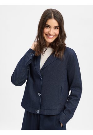 Uśmiechnięta kobieta o brązowych włosach do ramion nosi ciemnoniebieską zapinaną na guziki kurtkę na białej bluzce, pozując z jedną ręką przy głowie.