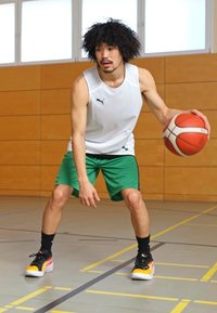 Sportininkas baltais be_rankovių marškiniais ir žaliais šortais, driblinguojantis krepšinio kamuolį. Juodi ir oranžiniai sportbačiai ant sporto salės grindų su žymėjimais.