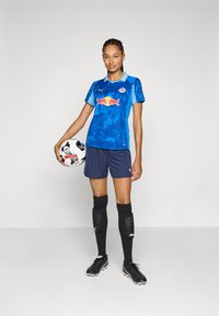 Maglietta da calcio blu con logo Red Bull, abbinata a pantaloni navy, calze al ginocchio nere e scarpe nere. Tiene in mano un pallone da calcio bianco e nero.