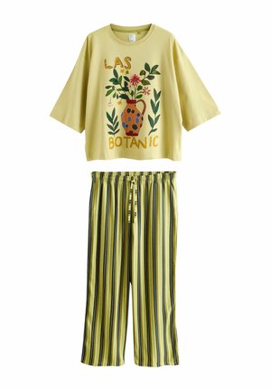 Gelbes Dreiviertelarm-Oberteil mit floralen Vasen-Print und dem Schriftzug "LAS BOTANIC", kombiniert mit gelben, schwarzen und grünen vertikal gestreiften Hosen.