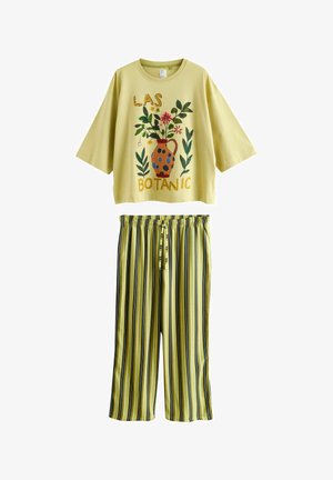 Kollane kolmeveerandvarrukaga top lillevaasi mustriga ja tekstiga "LAS BOTANIC", mida kombineeritakse kollaste, mustade ja roheliste vertikaalsete triibuliste pükstega.