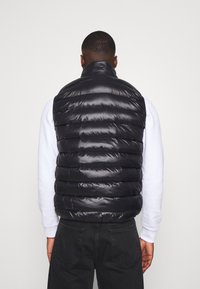 Schwarze gesteppte Weste mit hohem Kragen, mit einer wattierten Textur und glänzendem Finish. Wird über einem weißen Langarmshirt und dunklen Hosen getragen.