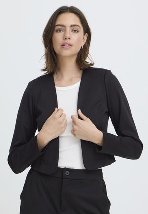Jonge vrouw in een zwart cropped blazer en broek met een witte geribbelde top, die haar blazer vooraan rechtzet tegen een effen lichte achtergrond.