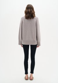 Masívny béžový pletený sveter s širokými rukávmi a rebrovaným lemom, v kombinácii s čiernymi slim-fit nohavicami a béžovými nazúvacími topánkami. Jednoduché biele pozadie.
