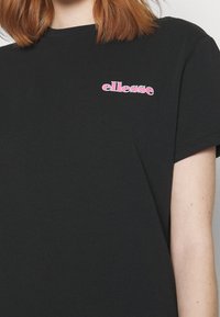 T-shirt en coton noir avec des manches courtes et un col ras du cou, arborant un logo "ellesse" rose imprimé sur la partie gauche de la poitrine.