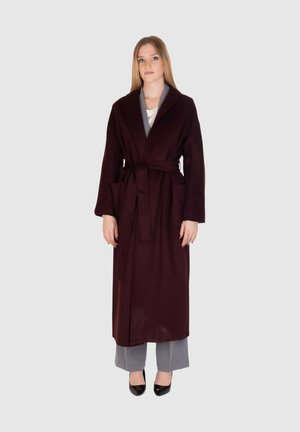 Lunga veste bordeaux con ampi rever, cintura in tessuto e tasche laterali; indossata sopra una maglia leggera e pantaloni grigi, caratterizzati da una trama liscia.