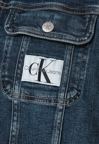 Detail na džínové bundě ukazující tmavě modrou látku, kontrastní švy, kovové patentní knoflíky a bílou štítek s černým textem: "Calvin Klein Jeans."