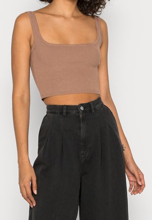 Femme portant un crop top côtelé marron et un pantalon noir ample taille haute sur un fond clair uni.
