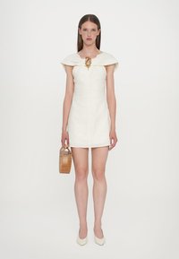 White linen mini dress with a cape collar, gold hardware detail, and fitted silhouette. Model holds a striped wooden bag and wears beige heels.