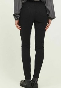 Los pantalones ajustados negros con una textura suave, que cuentan con dos bolsillos traseros y una cintura de altura media, combinados con una blusa gris fluida y zapatos negros.
