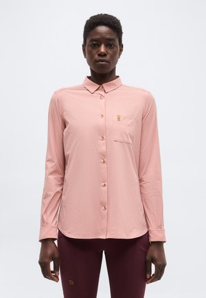 Άτομο που φοράει ανοιχτό ροζ πουκάμισο με κουμπιά και θήκη στο στήθος, συνδυασμένο με σκούρο βουργουνδί παντελόνι, στέκεται μπροστά σε μια απλή φόντο.