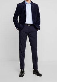 Marine corduroy blazer, lichtblauw shirt, op maat gemaakte donkere broek, en zwarte chelsea boots. De blazer heeft verticale ribbels en revers.