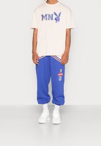 Person med en beige t-shirt med "MN" och en blå kaninlogga, blå mjukisbyxor med logotyper, vita strumpor och vita sneakers, stående.