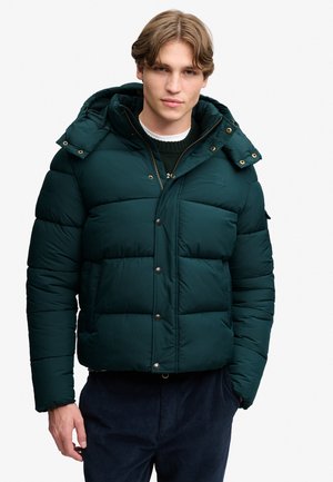 Joven con una chaqueta acolchada verde oscuro con capucha, suéter negro y pantalones oscuros, con una mano en el bolsillo sobre fondo blanco.