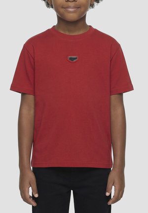 Bambino che indossa una maglietta rossa semplice a maniche corte con una piccola spilla nera e rossa al centro, abbinata a pantaloni neri, in piedi su sfondo grigio.