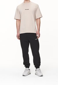 Mies, joka on pukeutunut beigeen lyhythihaiseen t-paitaan ja mustiin jogginghousuihin, molemmat merkitty "PEGADOR," valkoiset lenkkarit jalassa, seisoo tasaista valkoista taustaa vasten.