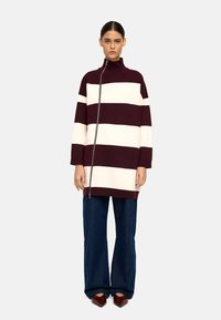 Przesadzony sweter z dzianiny w poziome bordowe i kremowe paski, z wysokim kołnierzem i bocznym zamkiem. Połączony z szerokimi ciemnoniebieskimi spodniami dżinsowymi.