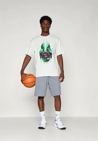 Hvit t-skjorte med kort erm og grønn og lilla grafisk design, parret med grå shorts, hvite joggesko og holder en basketball.