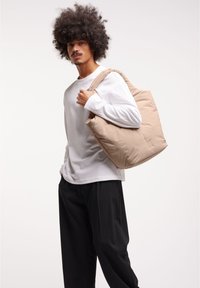 Borsa tote imbottita beige con finitura liscia, ampiezza delle cinghie e design minimalista, tenuta da una persona che indossa una camicia bianca a maniche lunghe.