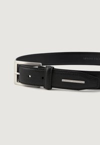 Mustaline nahast vöö, millel on hõbedane ristkülikukujuline kinnituse ja metallsilt Armani Exchange logoga vöö otsas, taustal ühevärviline taust.