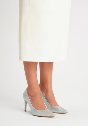 Szare skórzane czółenka na wysokim obcasie z ostrym noskiem i metalowymi akcentami, umieszczone na tle jasnej spódnicy.