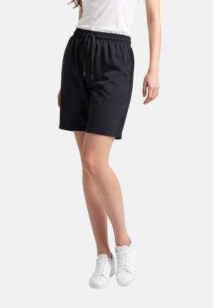 Donna con pantaloncini neri con coulisse, maglietta bianca e sneakers bianche, in piedi con le gambe incrociate su sfondo bianco.