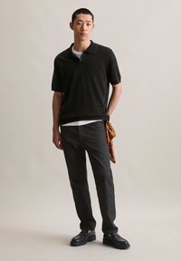 Młody mężczyzna ubrany w czarną koszulę polo z krótkim rękawem, czarne spodnie, czarne buty i brązową, wzorzystą chustkę włożoną w kieszeń, stojący na tle jednolitego tła.